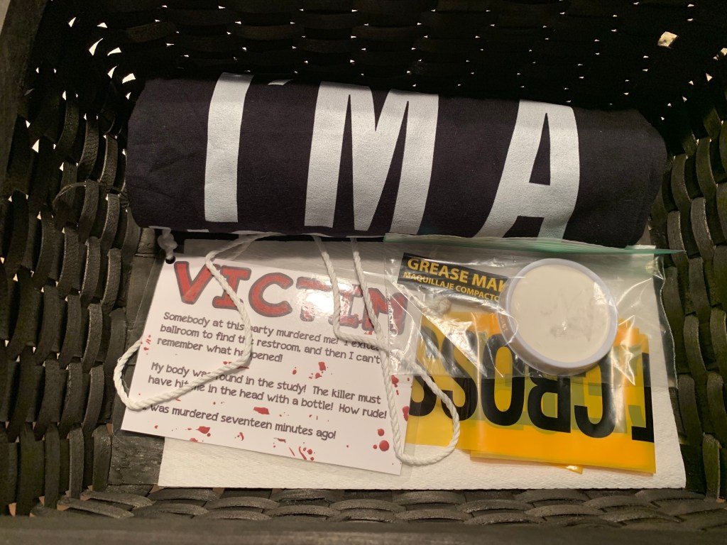 Close-up of ghost kit contents including a black “I’m a Ghost” shirt, victim sign, white face paint, crime scene tape, and instructions.