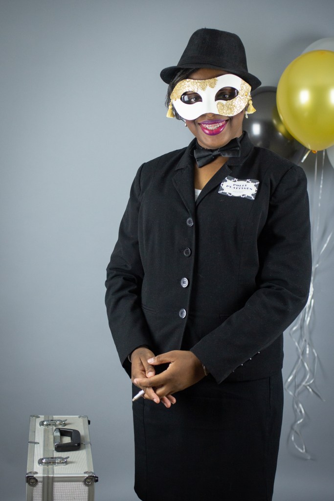 A woman in a black suit and hat poses with a gold and white masquerade mask. She holds a pen and stands next to a silver briefcase. Her name tag reads “Polly Plattygus"