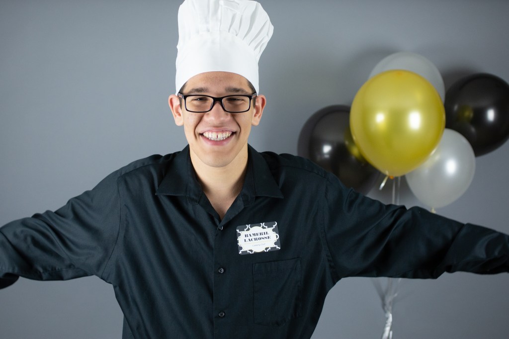 A smiling teen in a chef’s hat and black uniform stands with arms outstretched. He wears glasses and a name tag that reads “Bamarel Lacrosee - Celebrity Chef.”