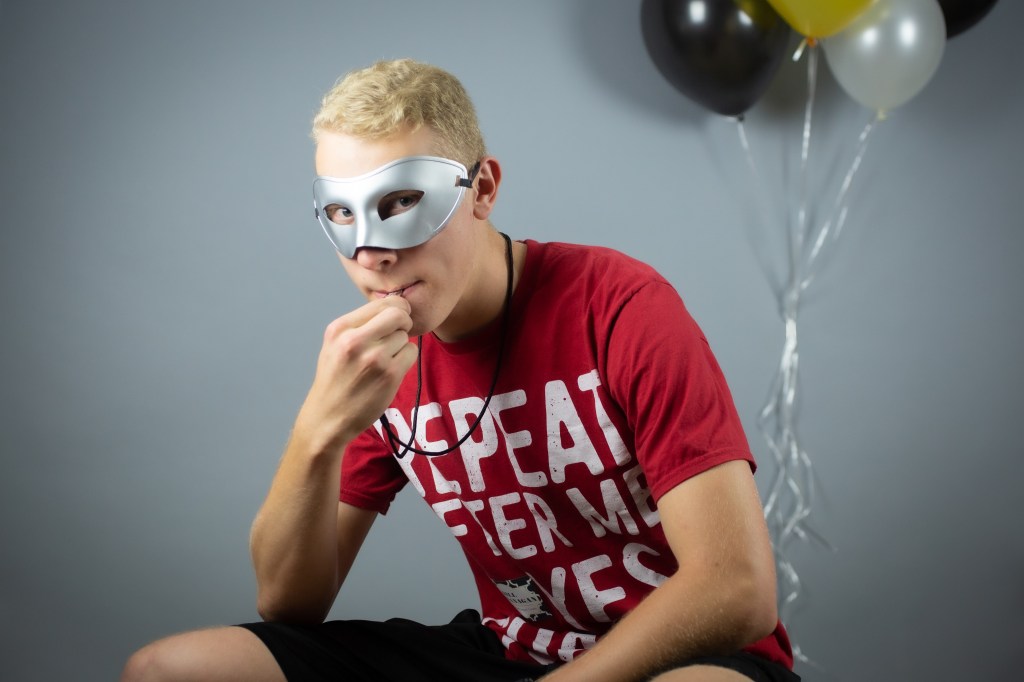 A masked teen in a red shirt blows a whistle and looks at the camera. He wears a silver masquerade mask.