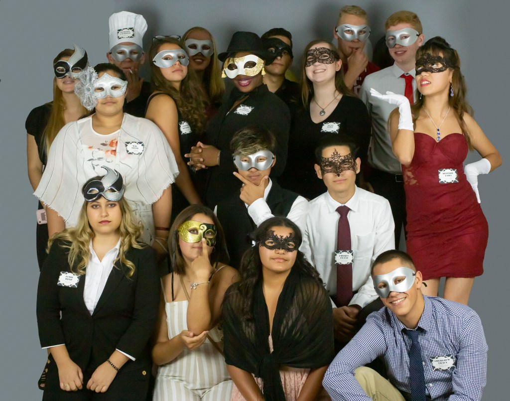 A group of teens in elegant and playful masquerade costumes pose together. Most wear decorative masks and formal or themed outfits. Characters include the chef, the masked agent, and Blondie Fawn.