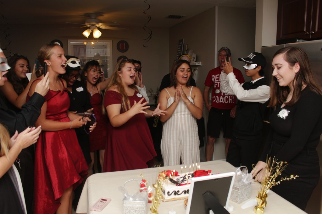 A group of teens in masquerade outfits gather around a murder-themed cake, laughing and singing as the birthday girl smiles.