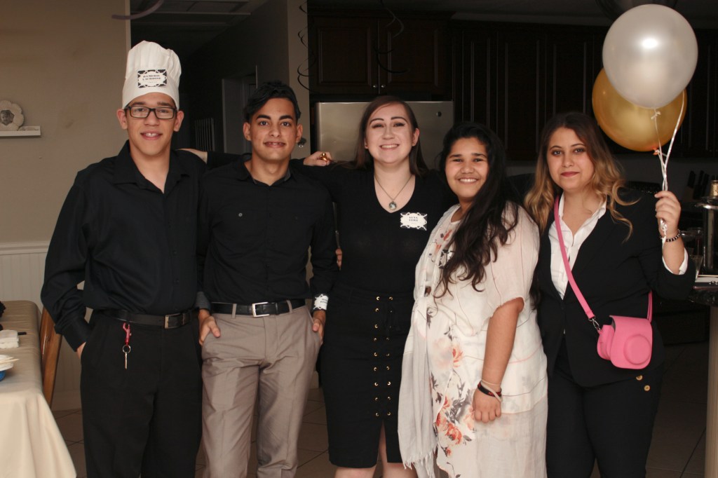 Five young adults pose together at the end of the murder mystery party, smiling in costume with one guest holding gold and white balloons.
