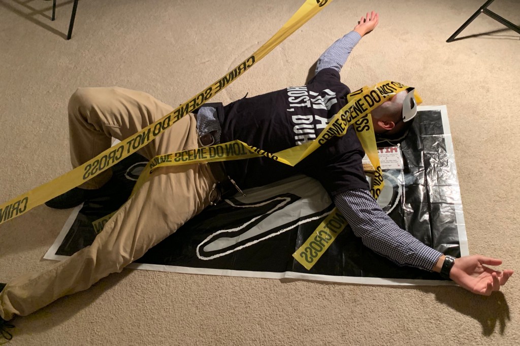 A close-up of a teen dressed as a murder victim wearing a black t-shirt, surrounded by yellow caution tape, lying across a printed body outline with a “victim” card nearby.