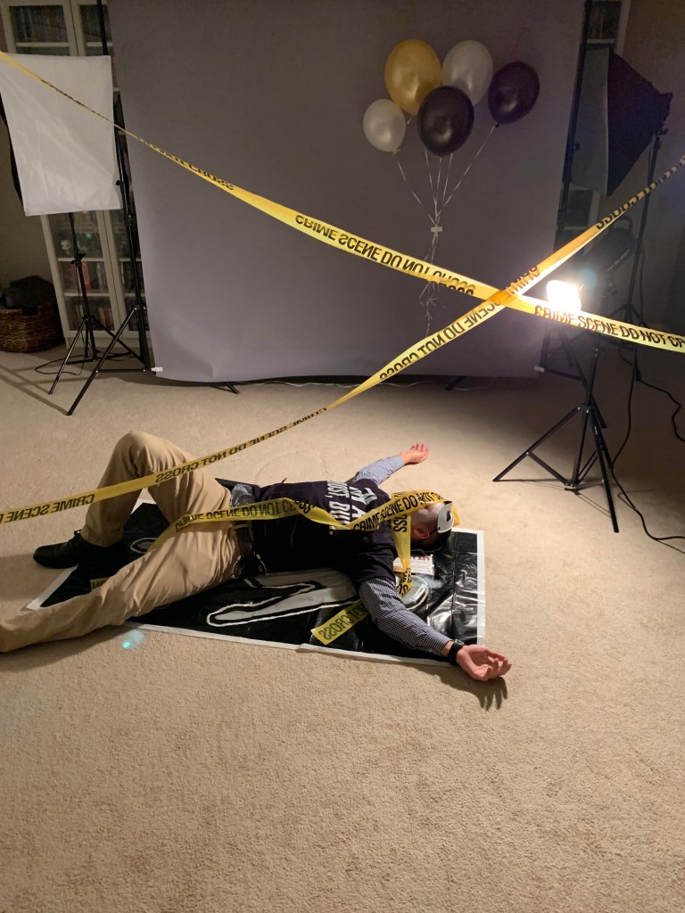 A wide shot of a fake murder victim staged at a crime scene with caution tape, balloons, dramatic lighting, and a backdrop. The victim wears a black “I’m a Ghost, Duh” shirt and a white mask.