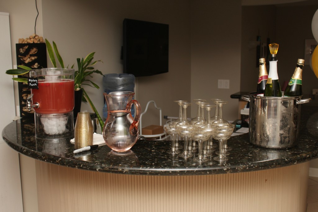 A bar setup with chilled bottles of sparkling cider, a dispenser of mystery punch with ice, stacked gold cups, a pink glass pitcher, and clear plastic goblets.