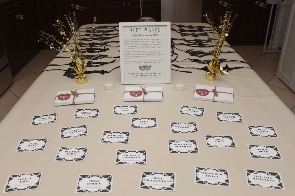 A full welcome table setup featuring a framed backstory sign, round packets tied with twine, printed name tags for each guest, masquerade masks, and gold star centerpieces.